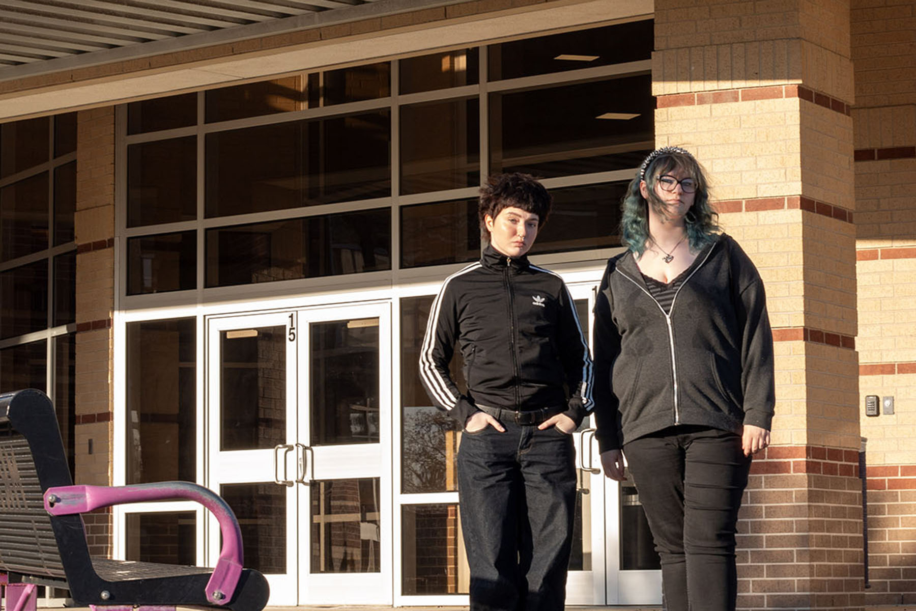 Two students stand outside a brick school building in warm sunlight.