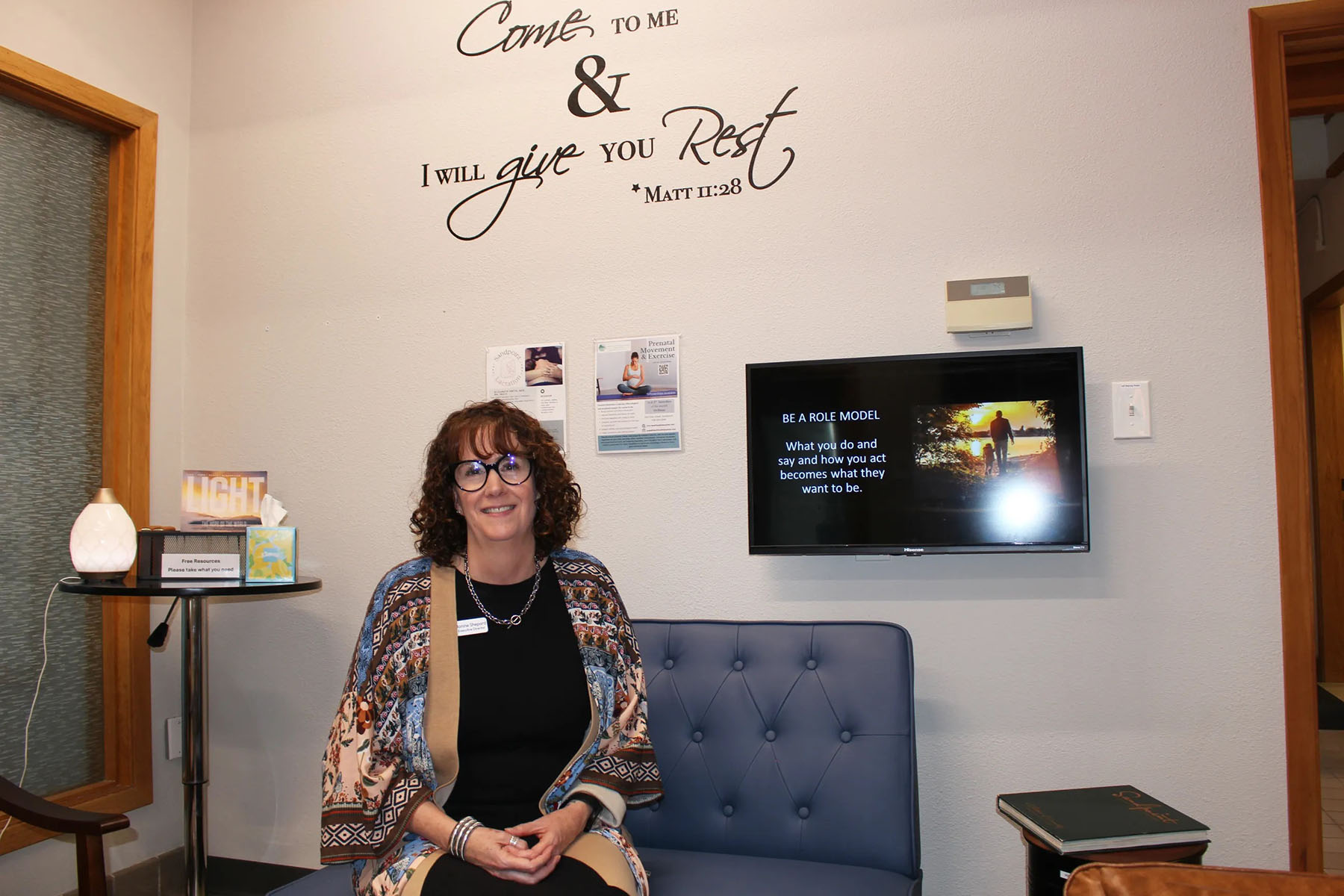 A woman sits on a blue bench in a care clinic waiting area beneath wall text reading, “Come to me & I will give you rest.”