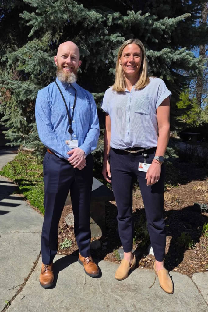 Two people stand outside in front of evergreen trees, wearing ID badges and smiling at the camera.