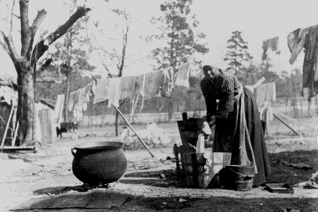 A Black woman washes clothes outdoors in a black and white historical photograph.