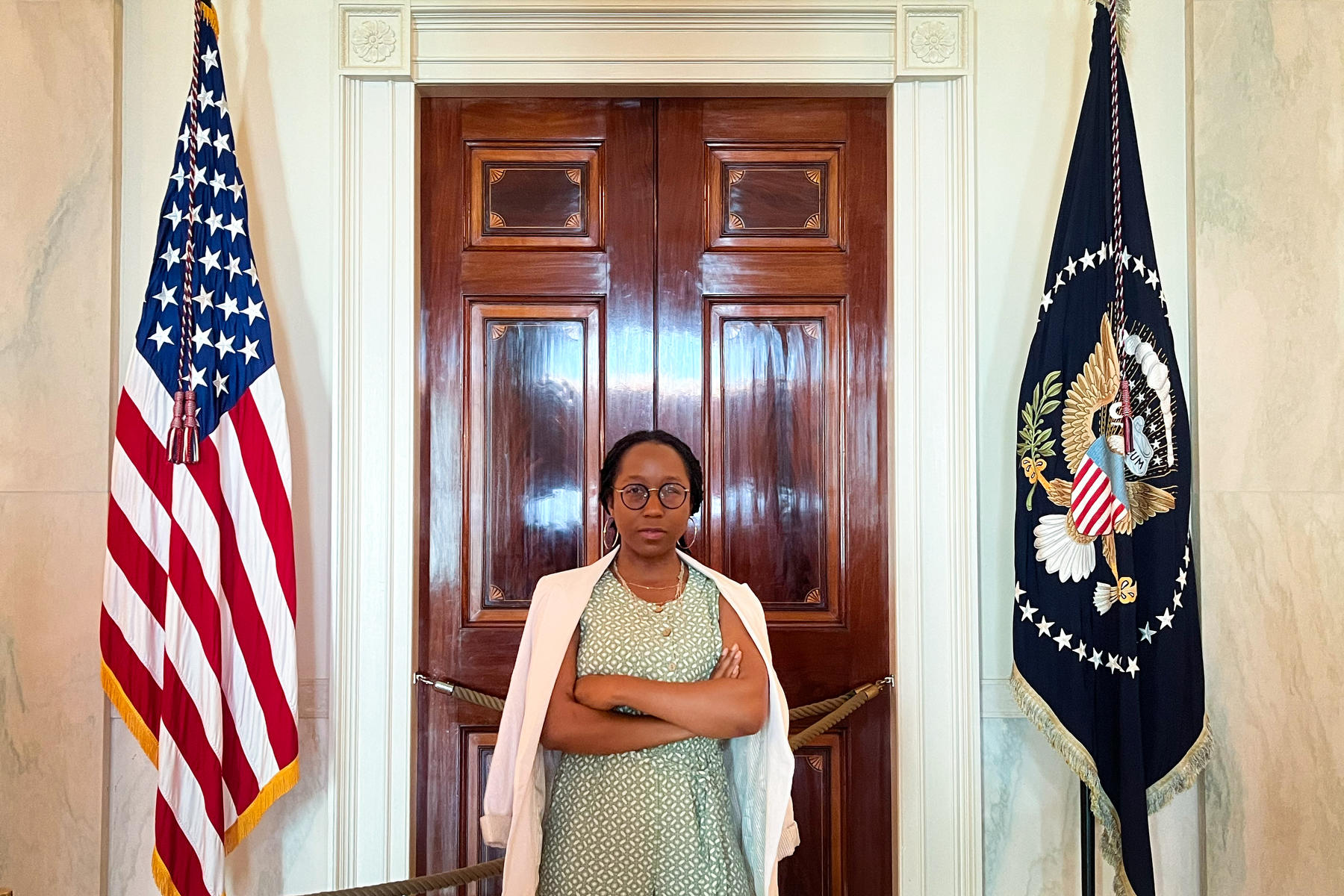 A woman stands in between two flags with her arms crossed.