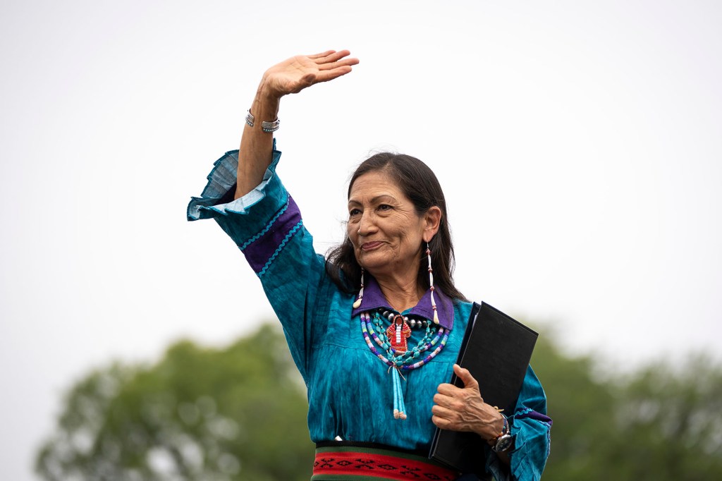 Deb Haaland in a turquoise blouse and beaded jewelry raises one hand while holding a black folder outdoors.