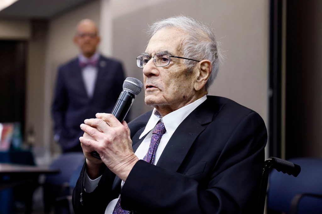 Barney Frank, sitting in a wheelchair and holding a microphone, speaks during an event.