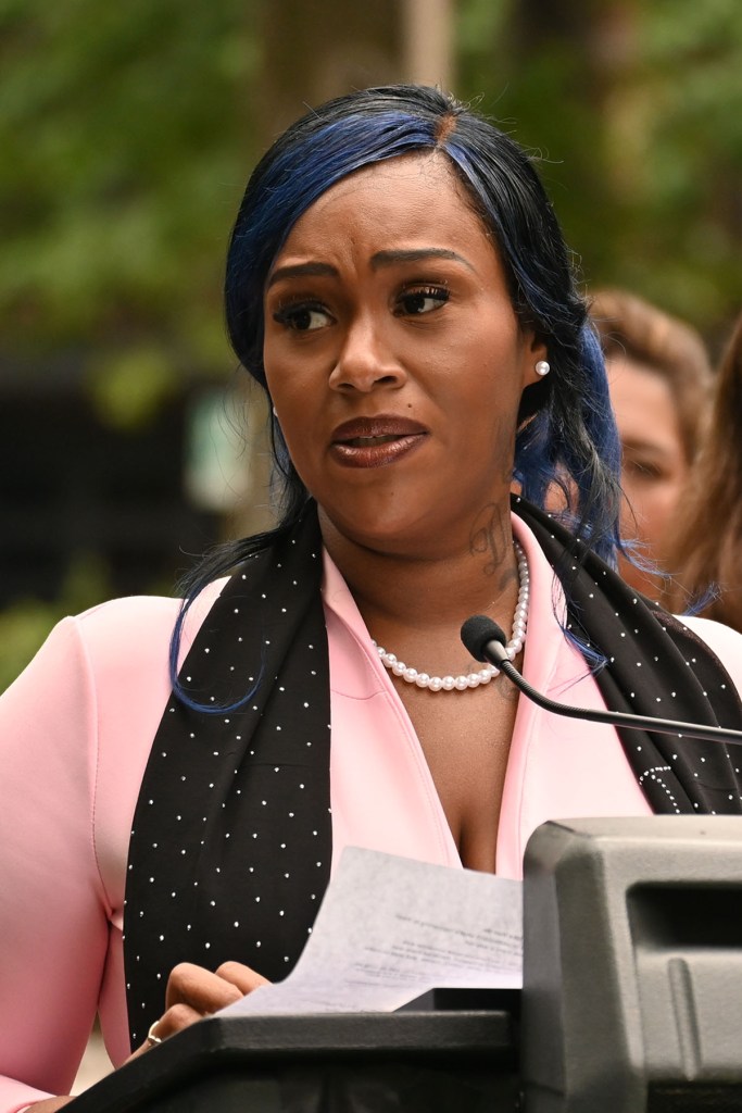 Jazz LedBetter stands at a podium outdoors, holding notes as she speaks. She wears a pink blazer , a black scarf, and a pearl necklace.