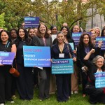 A large group of people poses outdoors on the lawn of the California state Capitol, holding blue signs that read “Save Lives” and “Protect Survivor Services.”