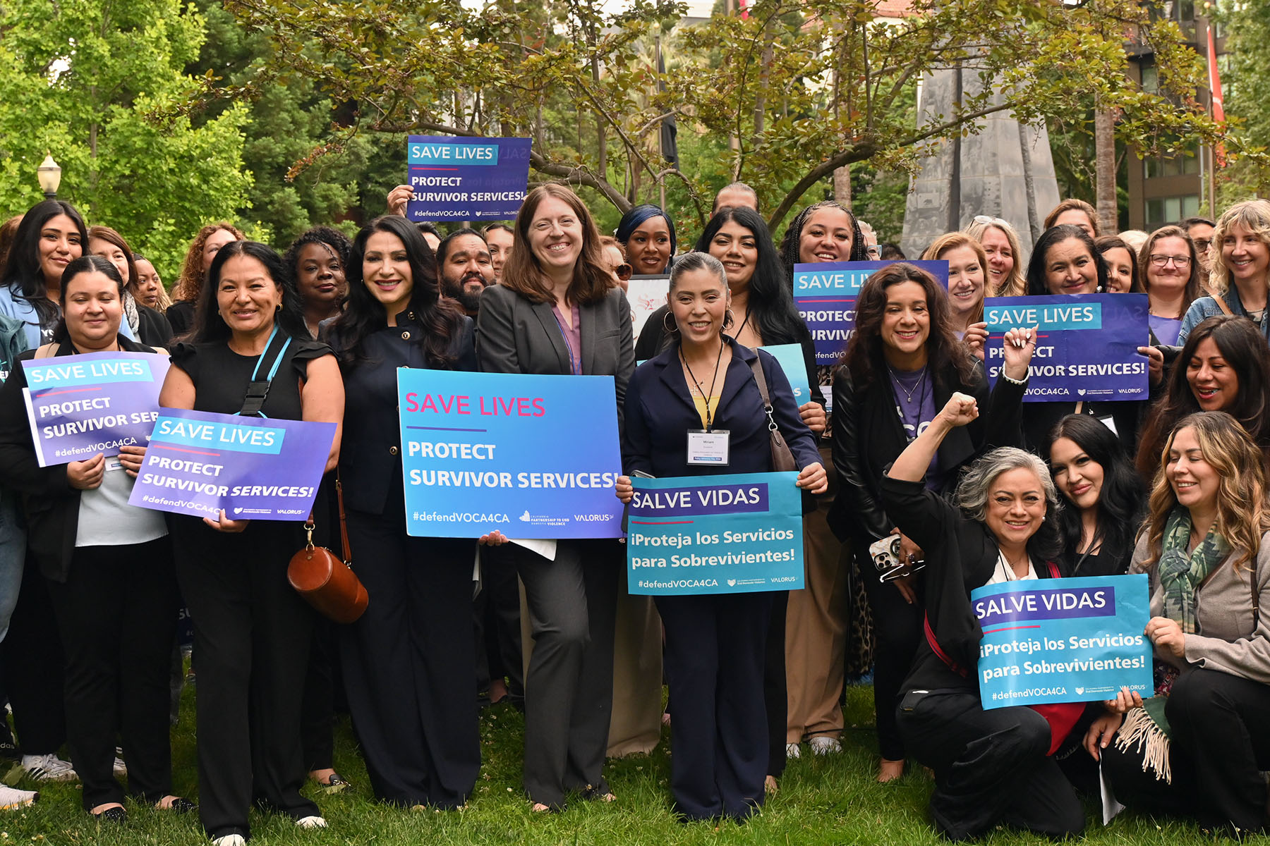 A large group of people poses outdoors on the lawn of the California state Capitol, holding blue signs that read “Save Lives” and “Protect Survivor Services.”