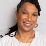 Kimberlé Crenshaw smiles at the camera while standing against a wall.