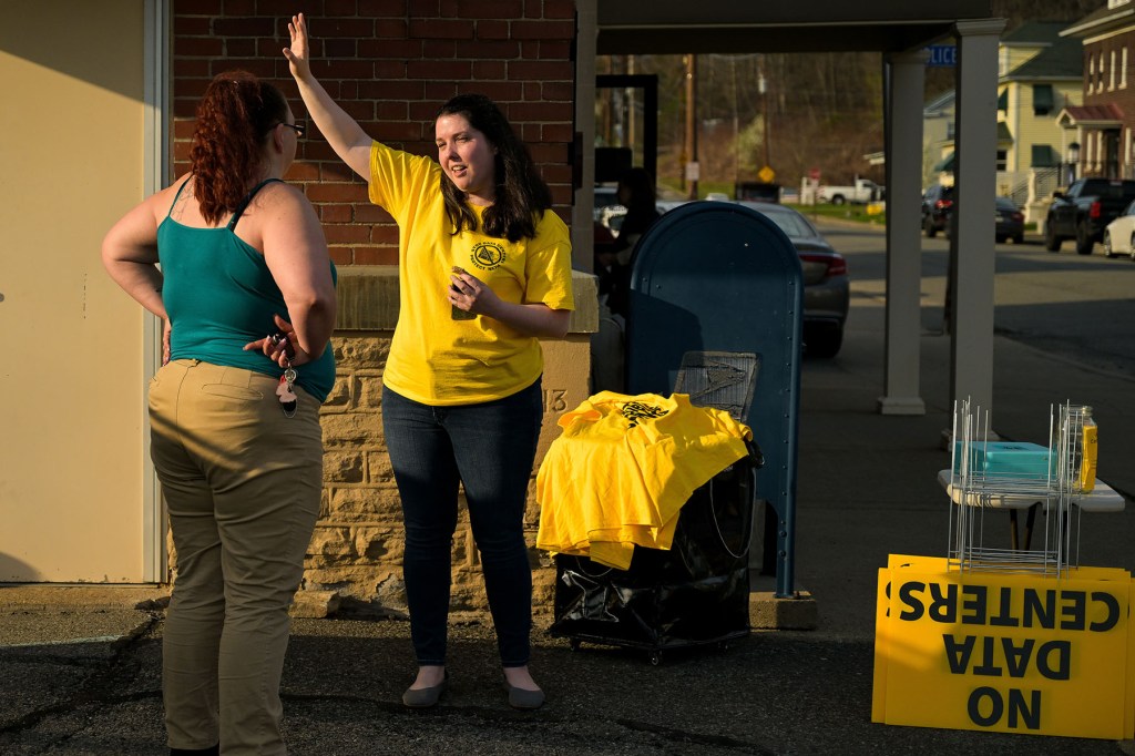 A woman in a yellow “No Data Centers” shirt talks with another woman outside a building, beside a table with flyers and yellow protest signs.