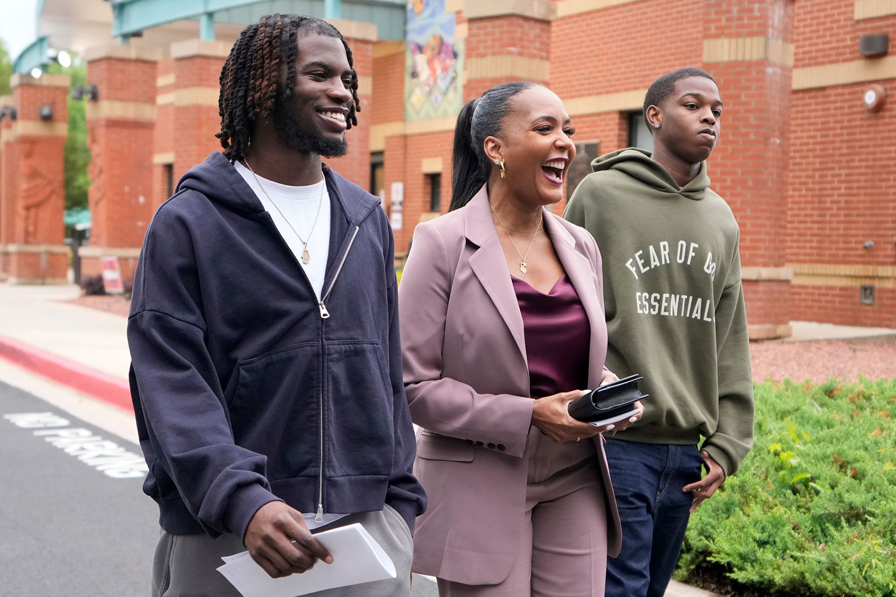 Three people walk outside a brick building. A woman in a mauve suit laughs while walking between two young men.