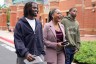 Three people walk outside a brick building. A woman in a mauve suit laughs while walking between two young men.