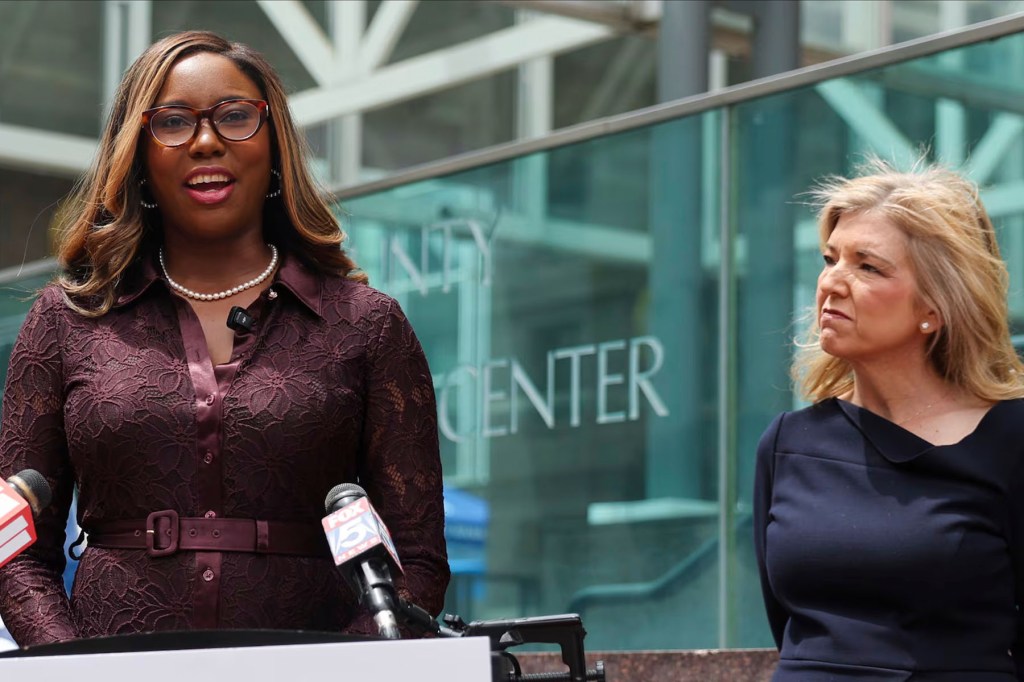 A woman speaks at a podium outside a glass-fronted building while another woman stands beside her and looks on.