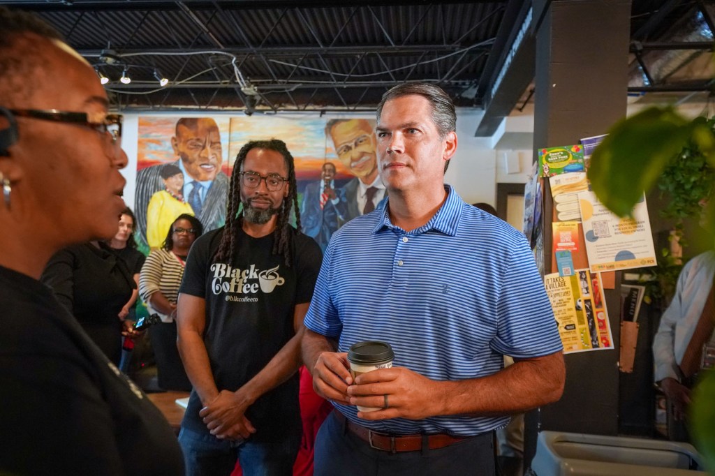 A man in a blue striped polo holds a coffee cup while speaking with people inside a coffee shop.