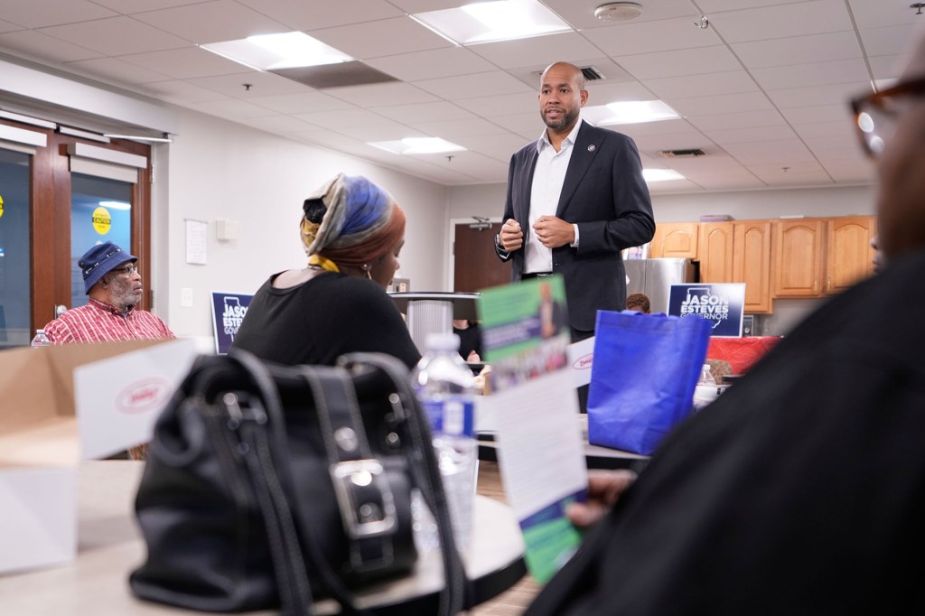 A man in a suit speaks to a group gathered around tables in a community room. Campaign signs that read “Jason Esteves Governor” are visible behind him.
