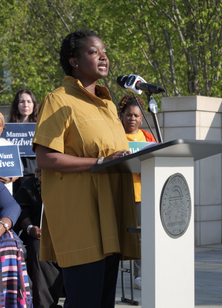 Tamara Taitt speaks at an outdoor podium in front of people holding signs about midwives and maternal care.