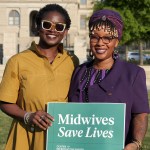 Tamara Taitt and Jamarah Amani smile while holding a green sign that reads “Midwives Save Lives.”