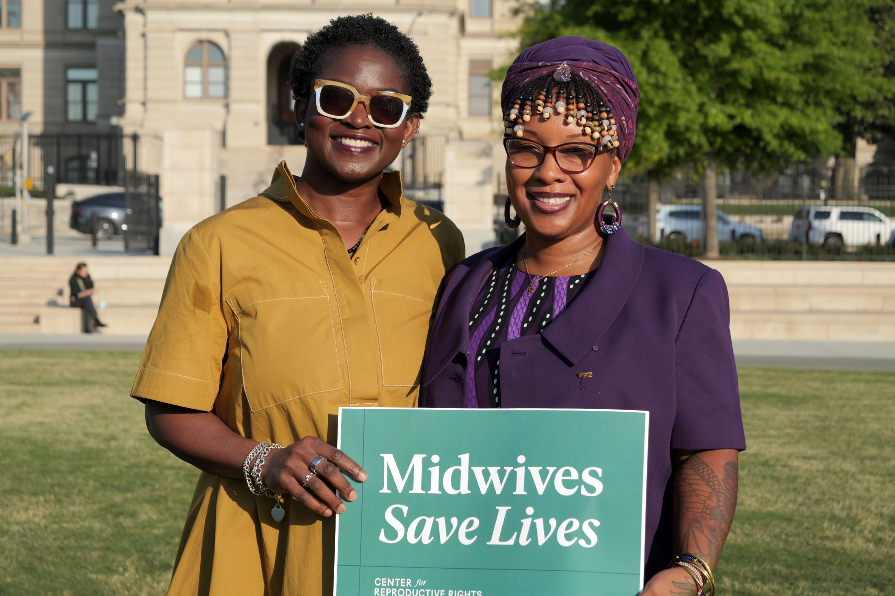 Tamara Taitt and Jamarah Amani smile while holding a green sign that reads “Midwives Save Lives.”