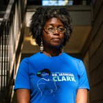 Dr. Jasmine Clark stands in a stairwell wearing glasses, patterned earrings and a bright blue campaign T-shirt that reads “re-elect Dr. Jasmine Clark.”