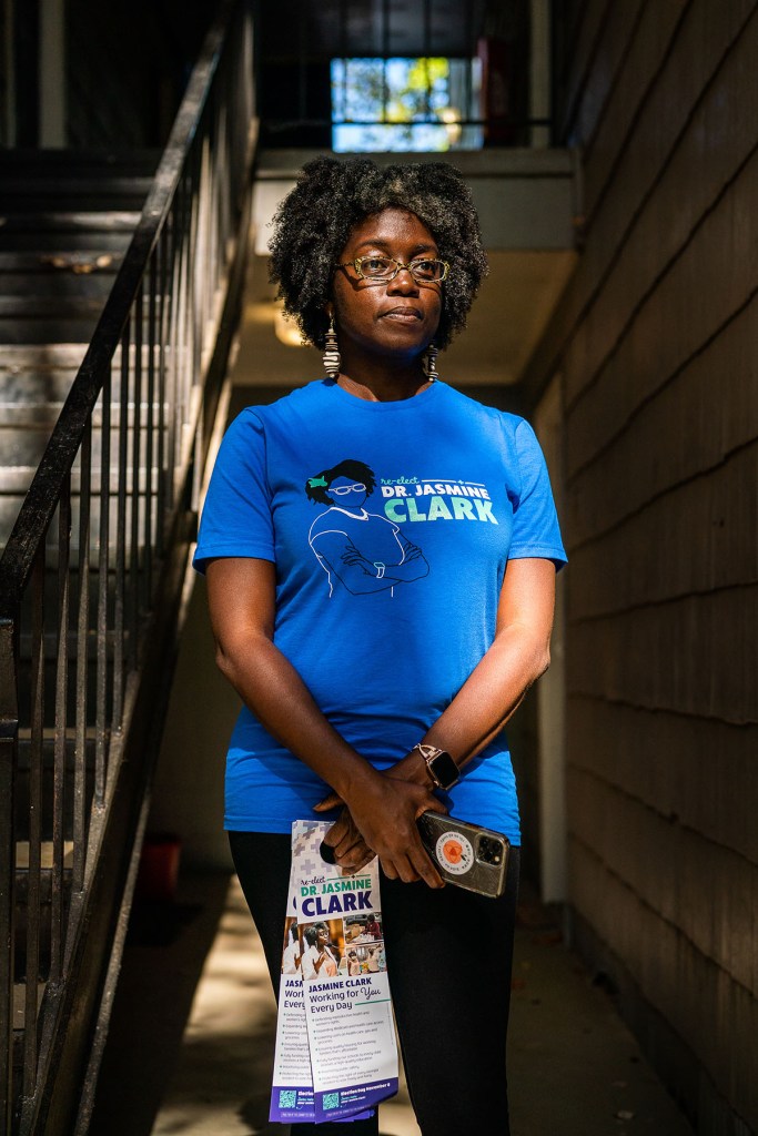 Dr. Jasmine Clark stands in a stairwell wearing glasses, patterned earrings and a bright blue campaign T-shirt that reads “re-elect Dr. Jasmine Clark.”