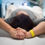 A patient lies on an exam table while an doula holds their hand during a procedure at a clinic.