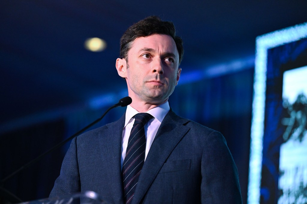Sen. Jon Ossoff stands at a podium in a dark suit and tie, looking off to the side under blue event lighting.