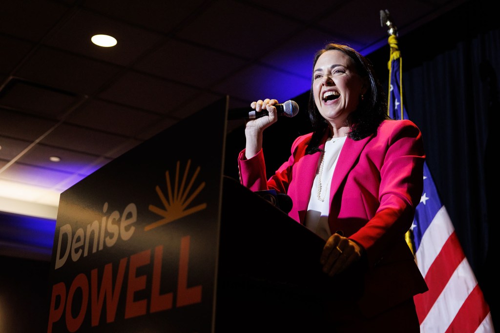 Denise Powell smiles while speaking into a handheld microphone at a campaign event. She wears a bright pink blazer and stands behind a podium with an American flag behind her.