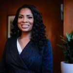 Precious Brady-Davis smiles while standing in an office with wood paneled walls and a potted plant near a window behind her.