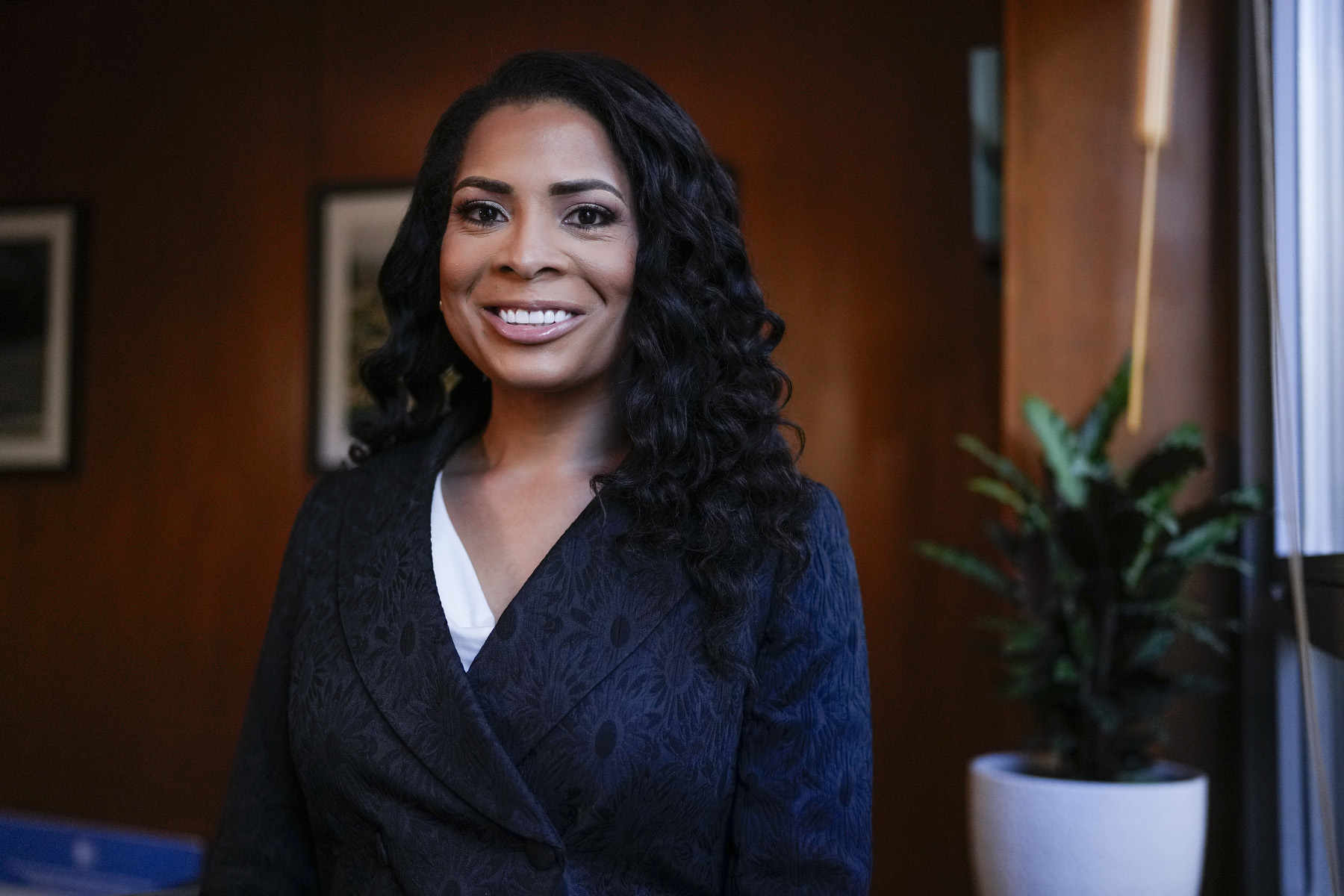 Precious Brady-Davis smiles while standing in an office with wood paneled walls and a potted plant near a window behind her.