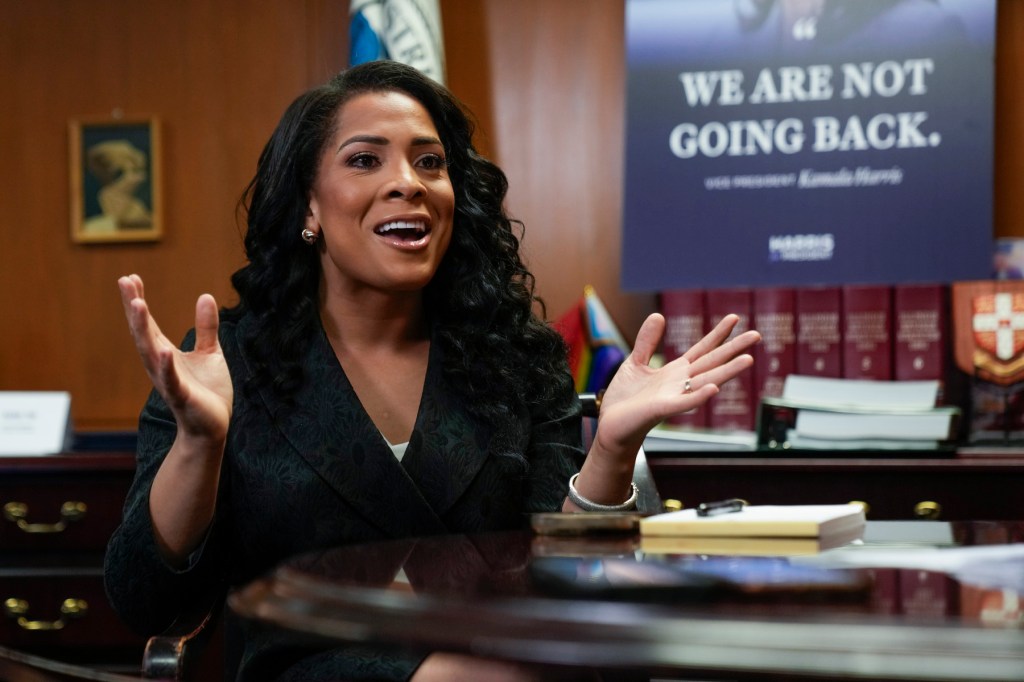 Precious Brady-Davis sits at a desk with her hands raised as she speaks. A sign behind her reads, “We are not going back.”