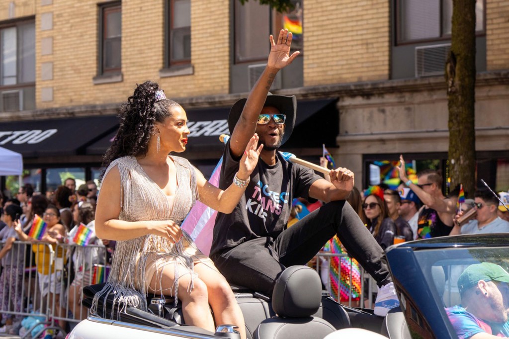 Precious Brady-Davis and Myles Brady-Davis sit on the back of a convertible and wave to a crowd at the Chicago Pride Parade. Pride flags are visible throughout the crowd behind them.