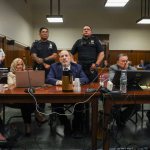 Several men and one woman sit at a table in a courtoom surrounded by police.