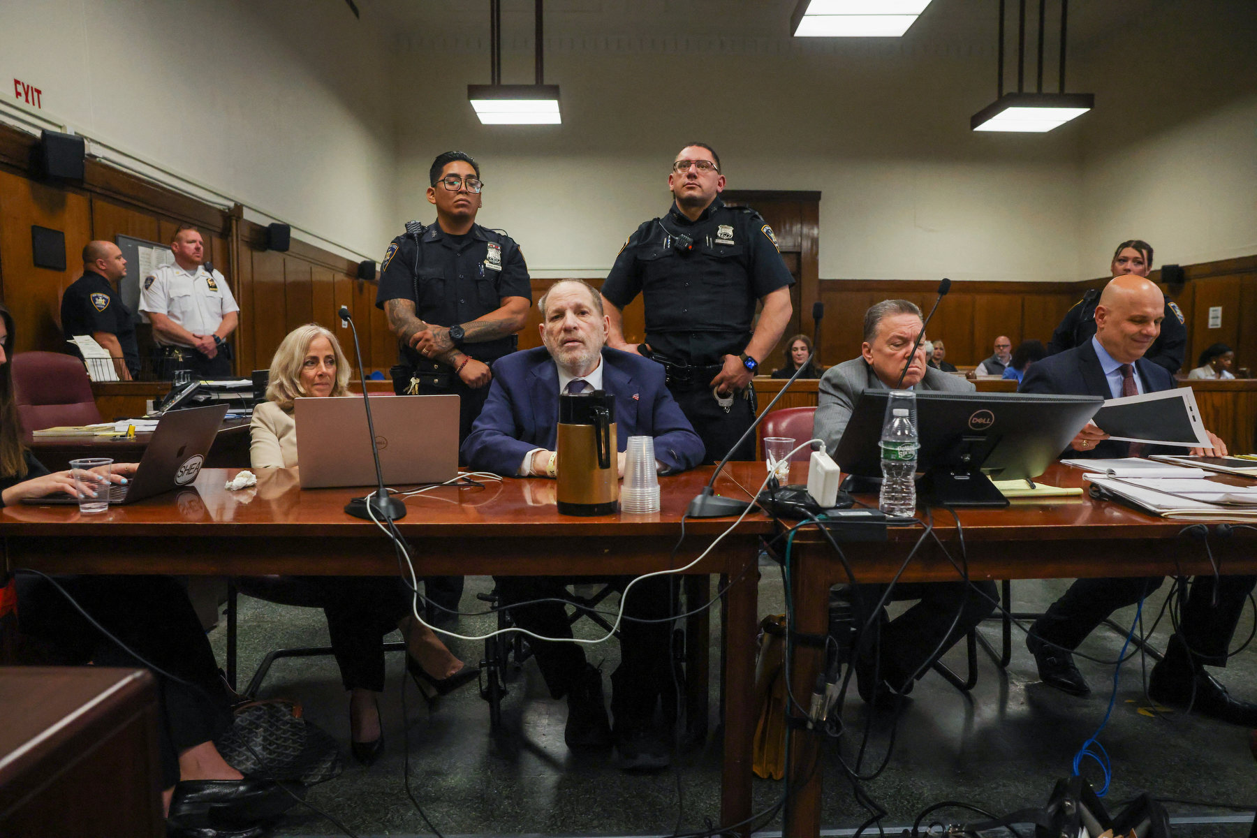 Several men and one woman sit at a table in a courtoom surrounded by police.