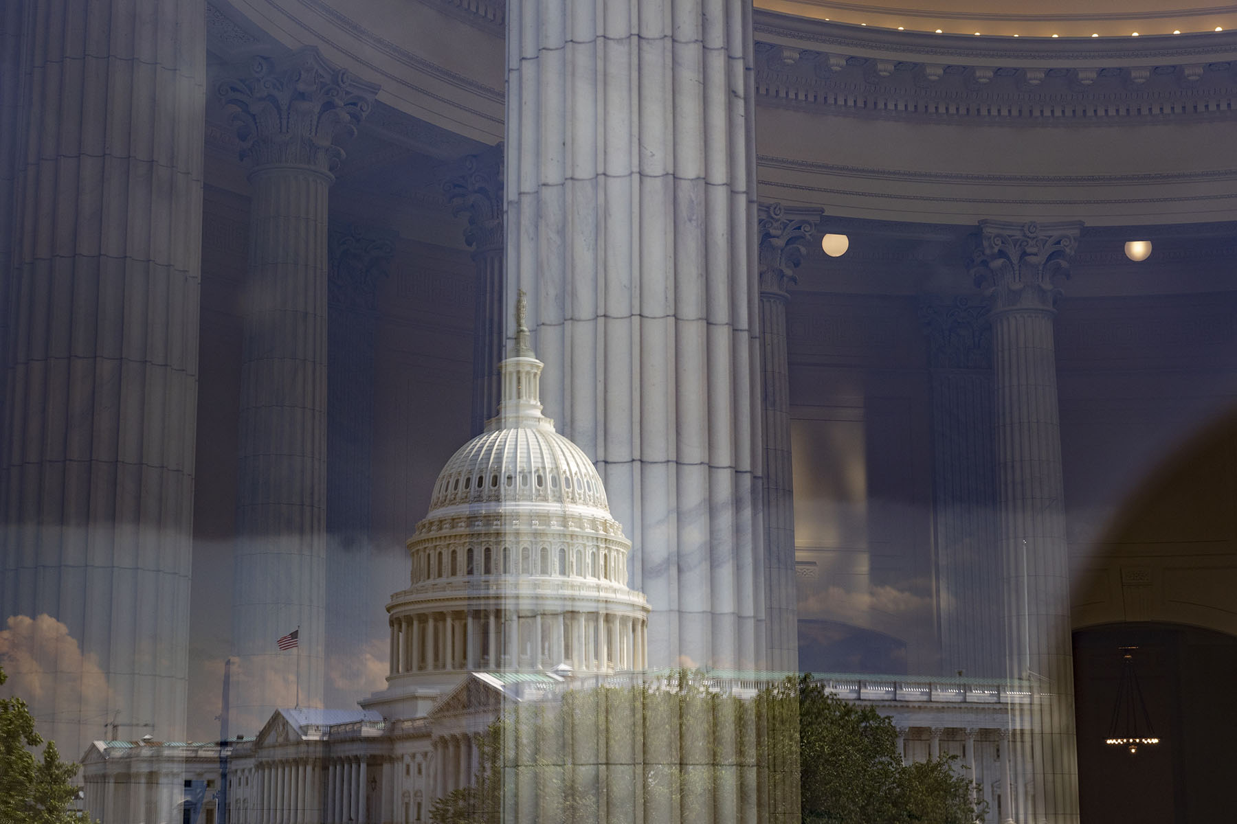 The Capitol dome is reflected over the interior of a building with tall marble columns.
