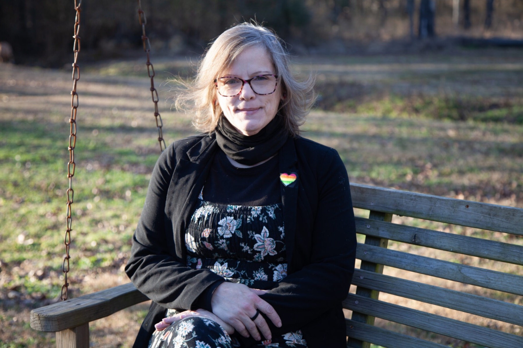 A woman in glasses sits on a wide wooden swing wearing a black blazer with a rainbow heart on the lapel.