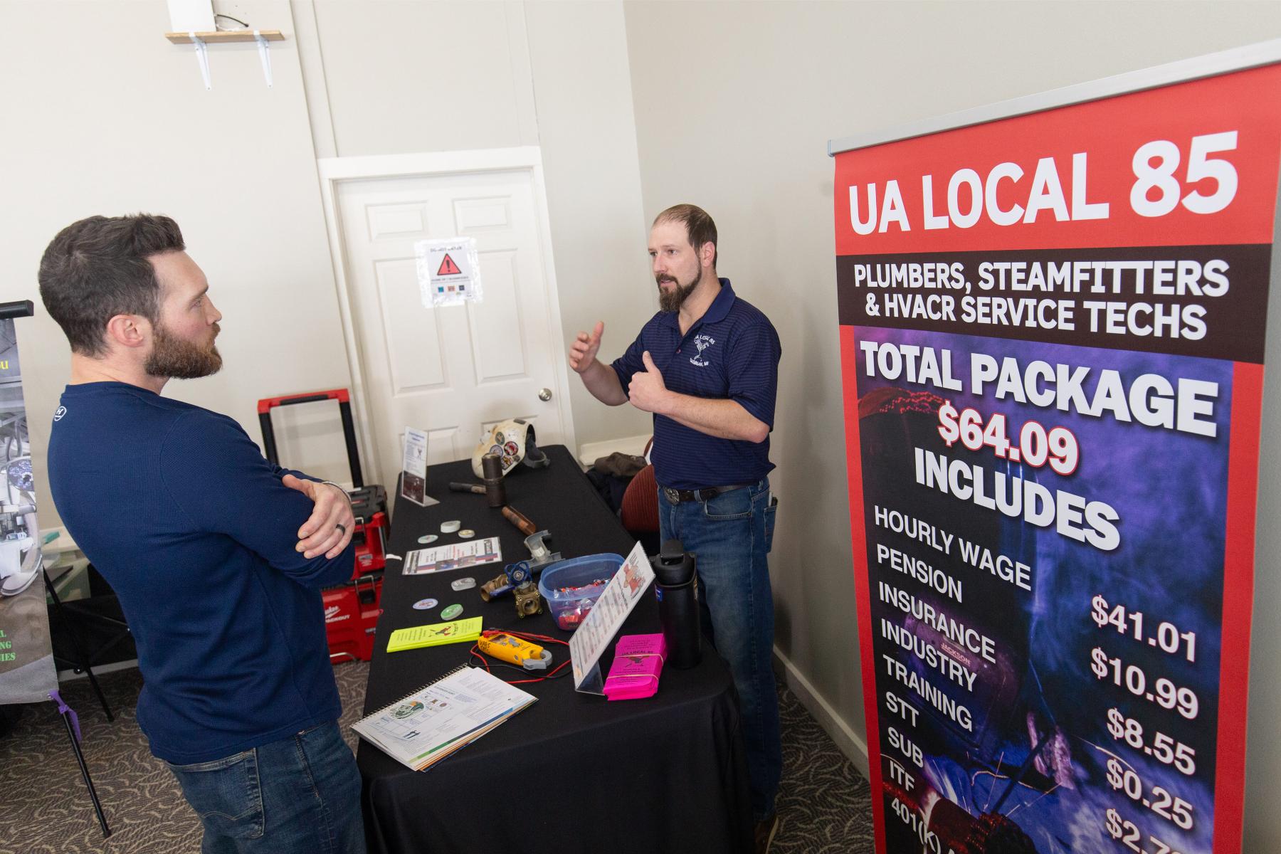 Plumbers union representative talks with a prospective trades student at a booth table