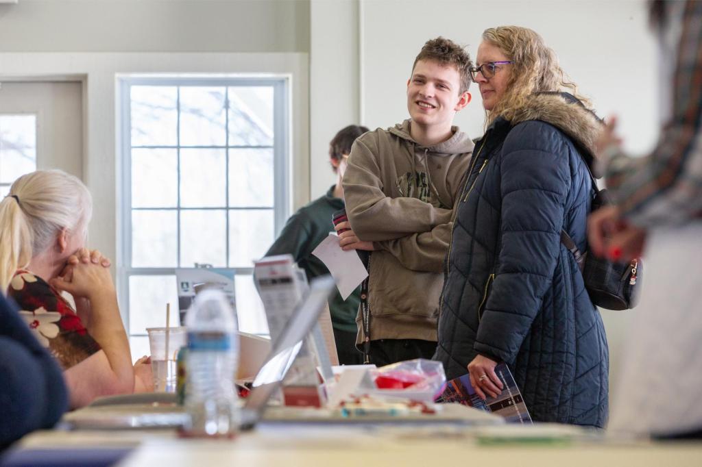 A smiling teen wearing a hoodie and a woman in a navy coat talk with a seated representative at a job fair table