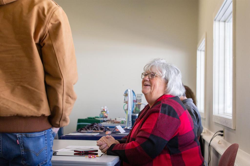 A woman in a red plaid sweater smiles up at a man in a tan work jacket across her job fair table