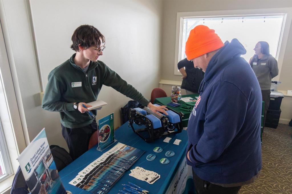 A recruiter explains a remotely operated vehicle to a man in an orange beanie at an education fair booth