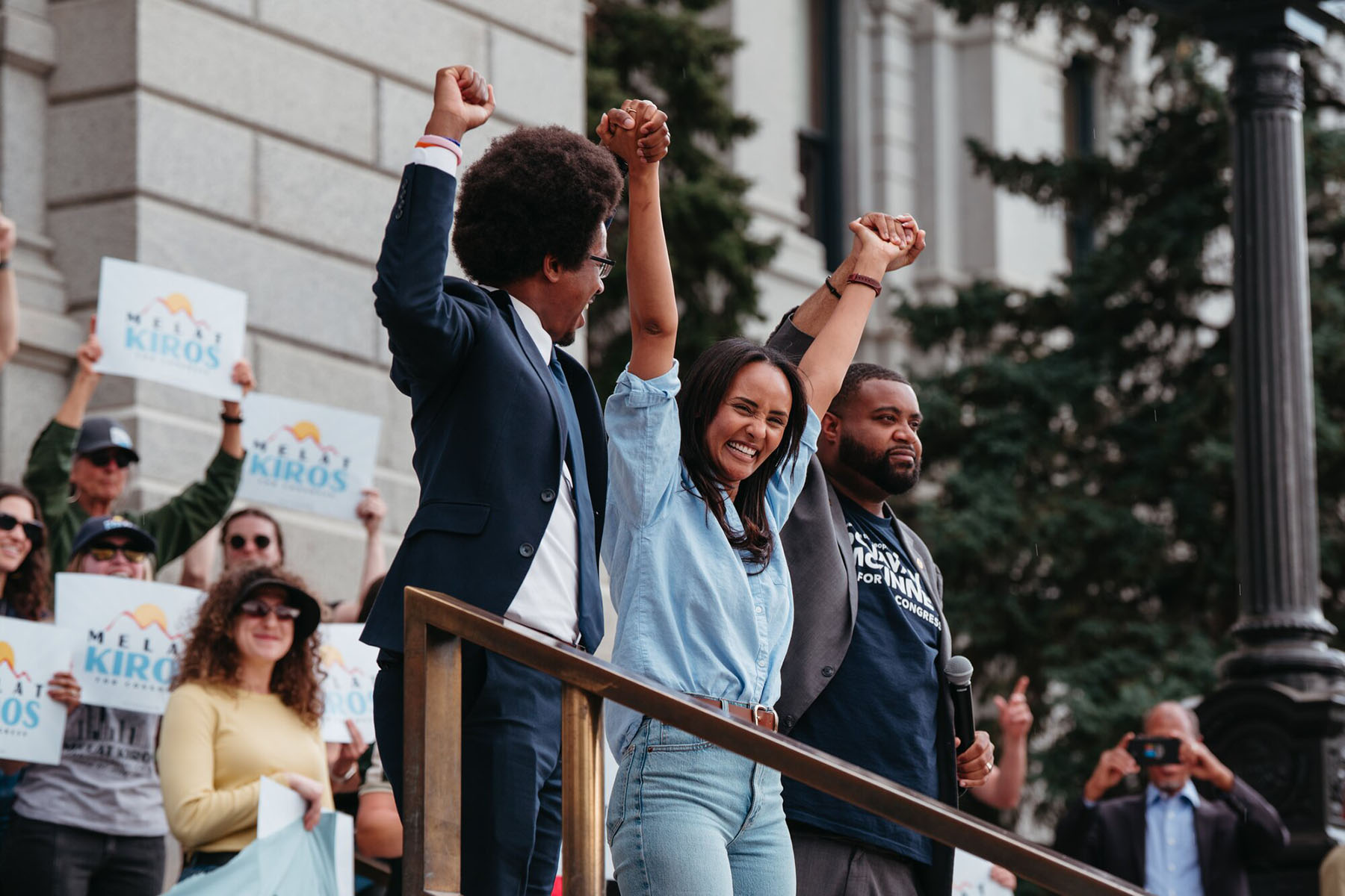 Melat Kiros stands on outdoor steps with two people, smiling as they hold their raised hands together. People behind them hold Melat Kiros campaign signs.
