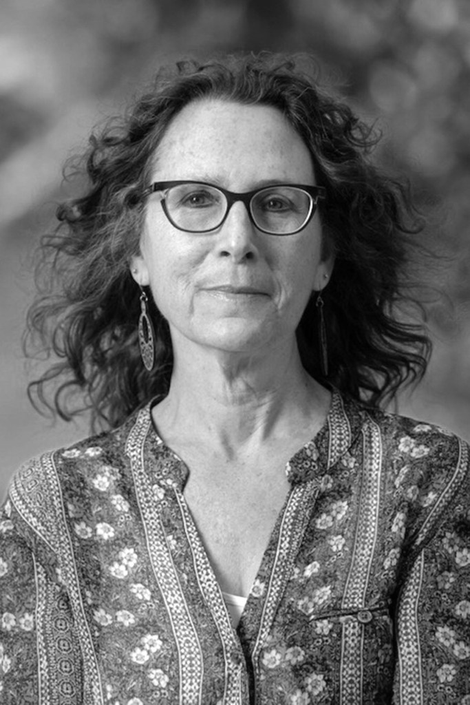 Headshot of Professor Julie Shayne looking at the camera and smiling softly. She is wearing dark rimmed glasses and a flowery button down.