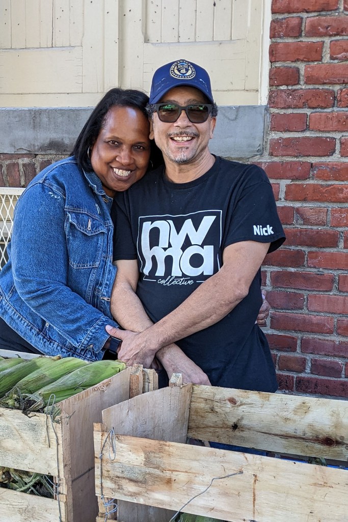 Linda James-Rivera and her husband, Nick, smile together behind wooden crates of corn at a community fridge event.