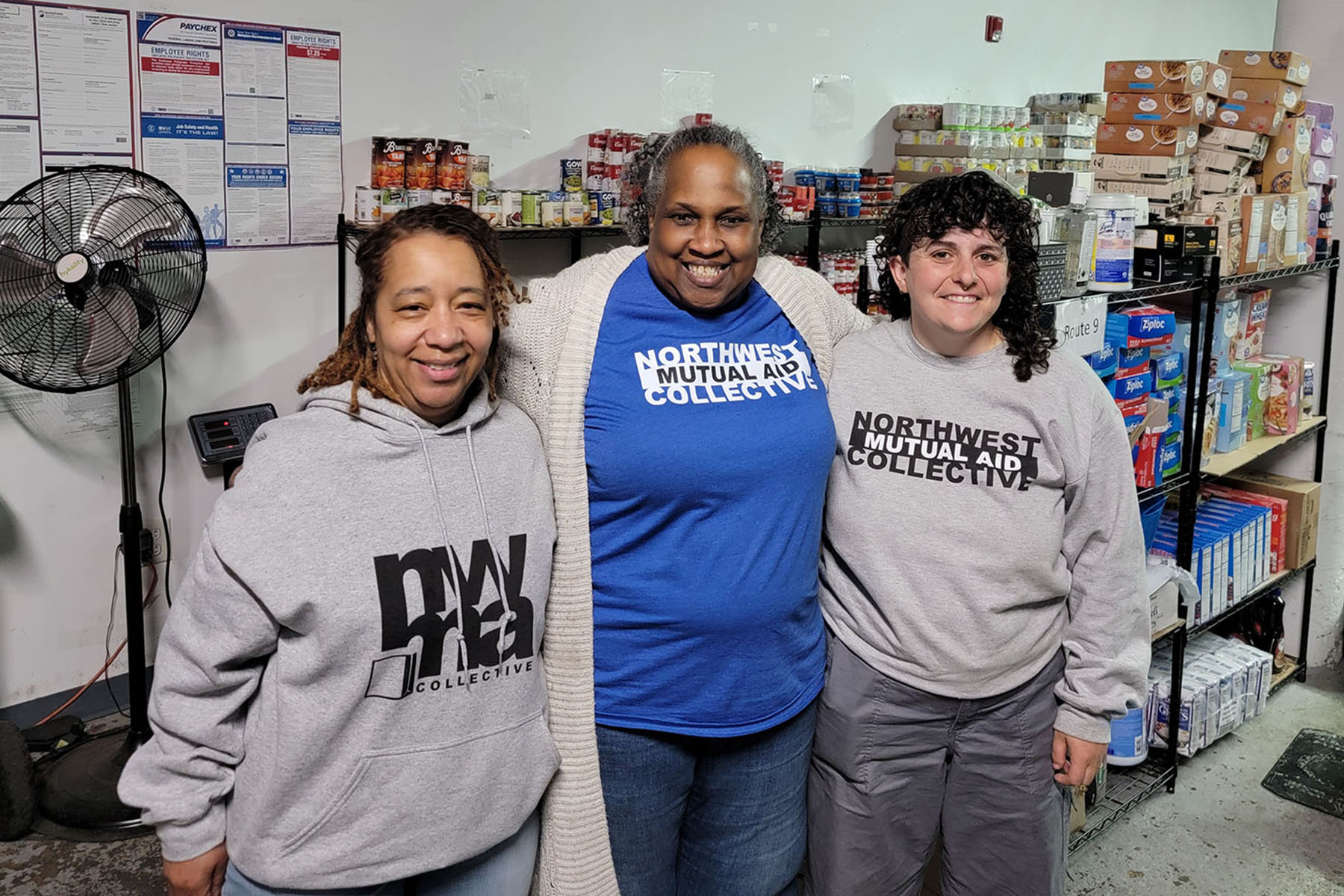 Renee Hill Joyner, Linda James-Rivera and Zoe Ganslaw stand together in front of shelves stocked with food and household supplies.