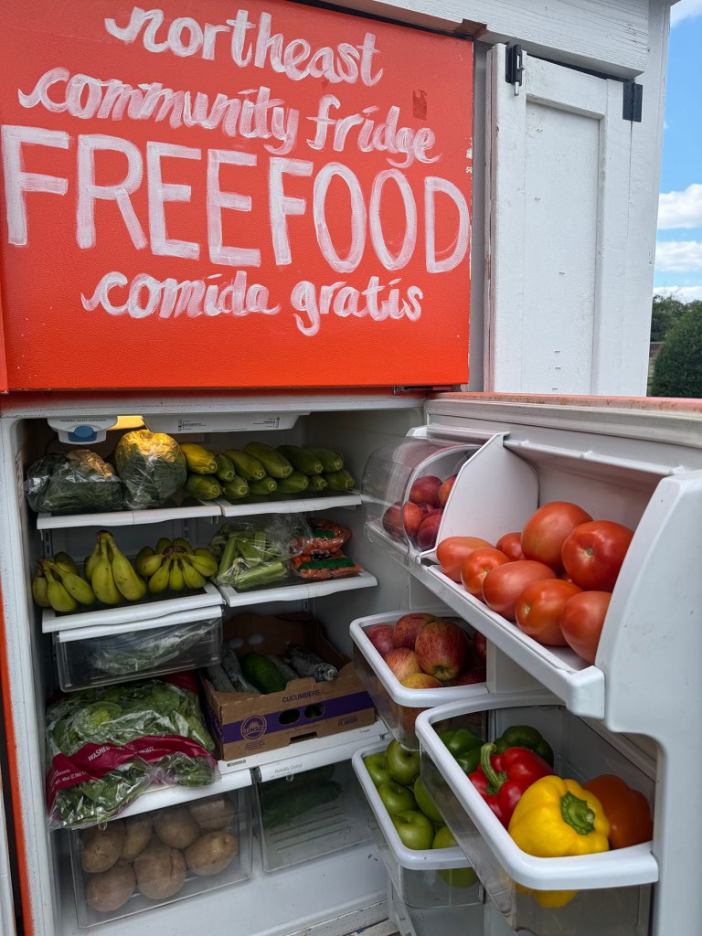 An open community fridge is stocked with fresh produce, including tomatoes, peppers, apples, cucumbers, bananas, lettuce and potatoes. A red sign above the fridge reads “Northeast Community Fridge,” “Free food” and “comida gratis.”