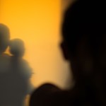 A silhouetted person stands in the foreground, with the shadow of an adult kissing a child on a warm yellow wall behind them.
