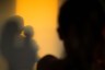 A silhouetted person stands in the foreground, with the shadow of an adult kissing a child on a warm yellow wall behind them.