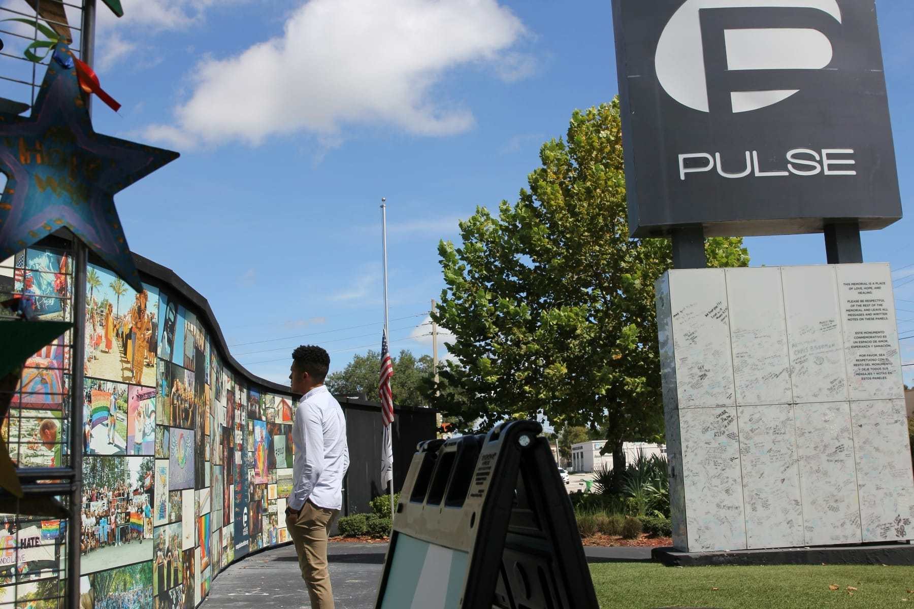 A large outdoor sign for Pulse nightclub is to the right, to the left is a wall with lots of images and a man standing looking at them
