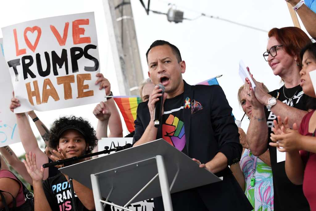 Carlos Guillermo Smith speaks into a microphone at a rally while someone nearby holds a "Love Trumps Hate" sign