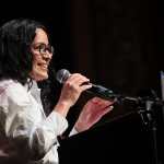 Claire Valdez speaks into a microphone; to the side is a sign that says 