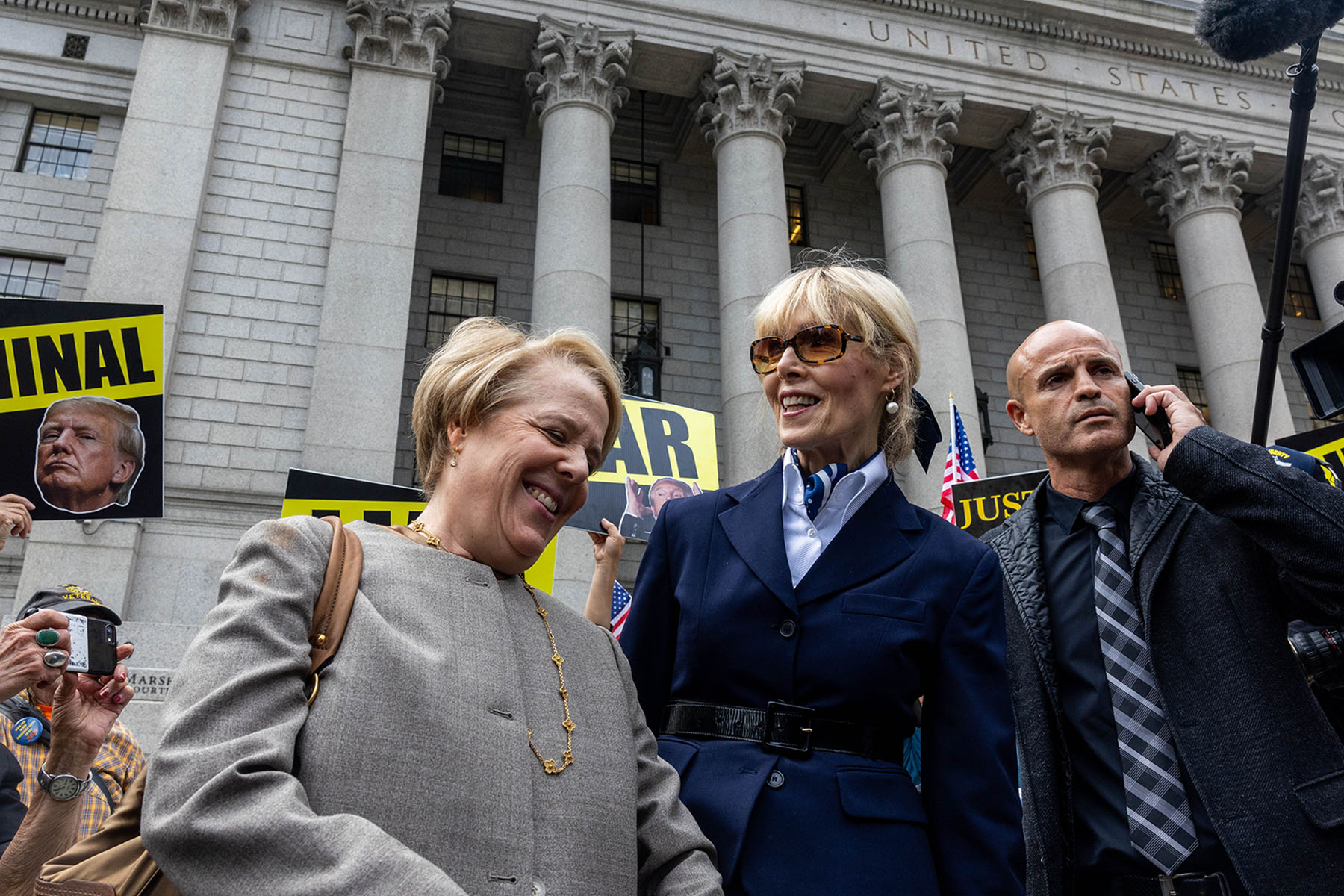 E. Jean Carroll walks outside a courthouse as demonstrators behind her hold signs criticizing Donald Trump.