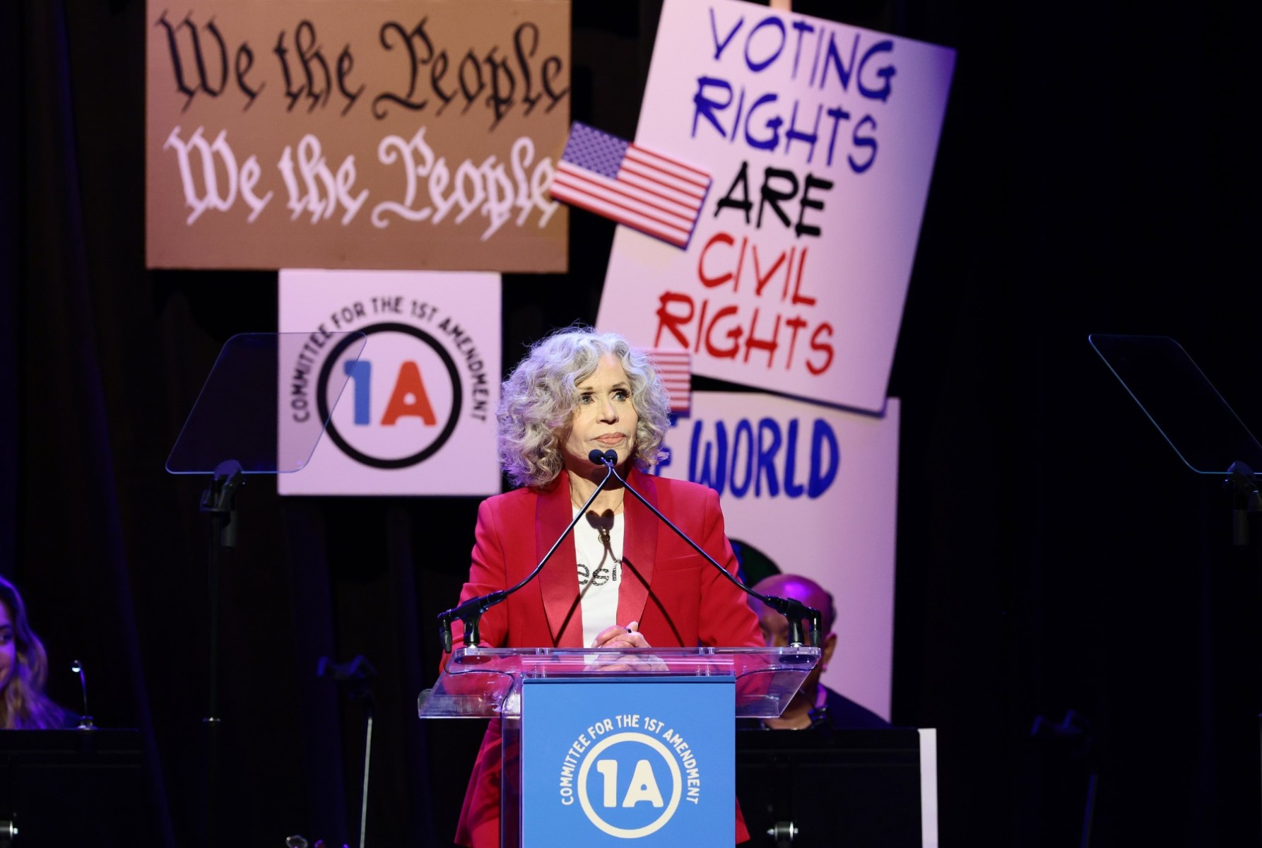 Jane Fonda, in a red suit, speaks onstage.