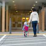 a woman holds the hand of a toddler who has a backpack on; they are seen from behind walking into a building